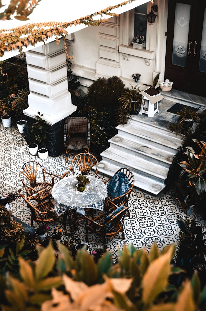 A cozy outdoor patio with wicker chairs and a tiled floor, surrounded by lush plants.